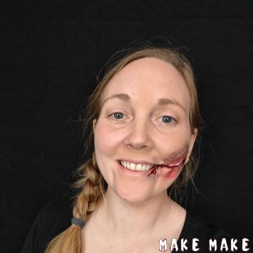 Smilende kvinde med stort kunstigt sr lavet med flydende latex fra mundvigen op langs kinden.
